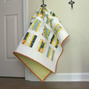 K&ouml;nnte beinhalten: Eine wei&szlig;e Steppdecke mit Patchwork-Muster, das florale und gestreifte Muster in Gelb-, Blau- und Gr&uuml;nt&ouml;nen zeigt. Die Steppdecke h&auml;ngt an einem Haken und hat einen gr&uuml;n-orangenen Rand.