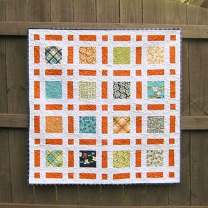 Może przedstawiać: Biała kołdra z kratką z pomarańczowych kwadratów i różnymi kolorowymi wzorzystymi kwadratami. Kołdra jest zawieszona na drewnianym płocie.