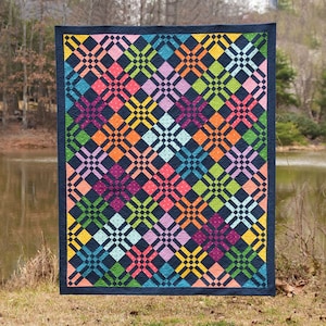May include: A colorful quilt with a geometric pattern of squares and diamonds. The quilt is made with a variety of bright colors, including pink, orange, yellow, green, blue, and purple. The quilt is displayed against a backdrop of a lake and trees.