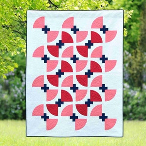 May include: A white quilt with a geometric pattern of pink and red half circles and navy blue plus signs.