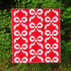 May include: A red quilt with a white bow pattern. The quilt is made of fabric squares sewn together to create a geometric design.