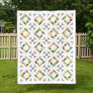 Pode incluir: Um edredom branco com um padrão geométrico de triângulos cinzentos e quadrados florais em tons de rosa, amarelo e verde. O edredom está pendurado ao ar livre em frente a uma cerca branca.