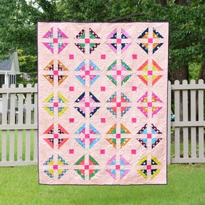 May include: A colorful quilt with a light pink background and a repeating pattern of squares and triangles in various colors. The quilt is hanging outside on a sunny day, with a white picket fence and green grass in the background.