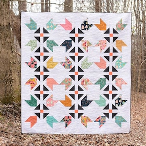 May include: A white quilt with a geometric pattern of black triangles and colourful squares. The quilt is made up of a variety of fabrics with floral and geometric designs. The quilt is hanging on a tree in a forest.