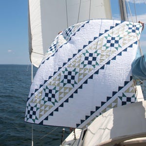 Może przedstawiać: Biała kołdra z geometrycznym wzorem w odcieniach niebieskiego i zielonego. Kołdra jest trzymana na żaglówce.