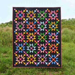 May include: A colorful quilt with a black background and a repeating pattern of star-like shapes in pink, blue, green, orange, and yellow. The quilt is bordered with a thin pink edge and is displayed outdoors against a backdrop of green grass and a cloudy sky.