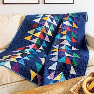 May include: A navy blue quilt with a geometric design of colorful triangles in shades of yellow, orange, red, green, purple, and white. The quilt is draped over a cream-colored sofa, showcasing its intricate pattern and texture.