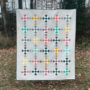 Puede incluir: Una colcha blanca con un diseño de patchwork que presenta cuadrados en varios colores, incluyendo rosa, verde azulado, amarillo y gris. La colcha está colgada de un tendedero en una zona cubierta de hierba.