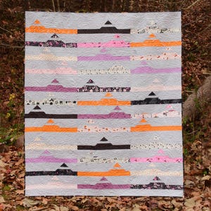 Könnte beinhalten: Eine handgefertigte Steppdecke mit grauem Hintergrund und horizontalen Farbstreifen. Die Streifen weisen orange, schwarze, rosa und violette geometrische Formen auf. Die Steppdecke wird im Freien mit einem Hintergrund aus Bäumen und Herbstlaub ausgestellt.