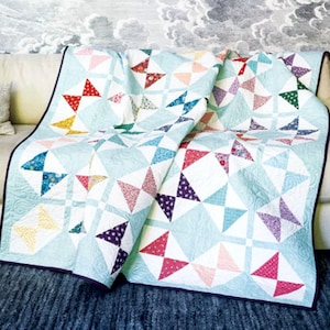 May include: A light blue quilt with a geometric star pattern, draped over a beige sofa. The quilt features colourful triangles in red, pink, yellow, blue, and purple. The quilt is bordered with a dark blue binding.