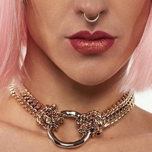 May include: Gold-toned chain necklace with a large silver-colored ring pendant. The necklace features a thick chain design with intricate detailing around the ring. The image shows a close-up view of the necklace against a skin tone background.