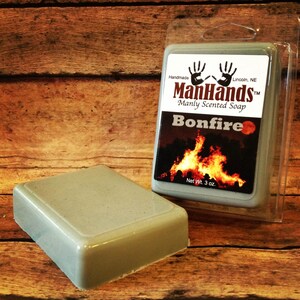 May include: A grey bar of soap with the text "ManHands™ Manly Scented Soap Bonfire" and a picture of a campfire. The soap is packaged in a clear plastic container. The soap weighs 3 ounces.