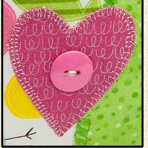 Pode incluir: Um cora&ccedil;&atilde;o de tecido rosa com bordas costuradas brancas e um bot&atilde;o rosa no centro. O cora&ccedil;&atilde;o &eacute; decorado com redemoinhos brancos e assenta sobre um fundo padronizado com detalhes verdes e amarelos. O design &eacute; alegre e artesanal.