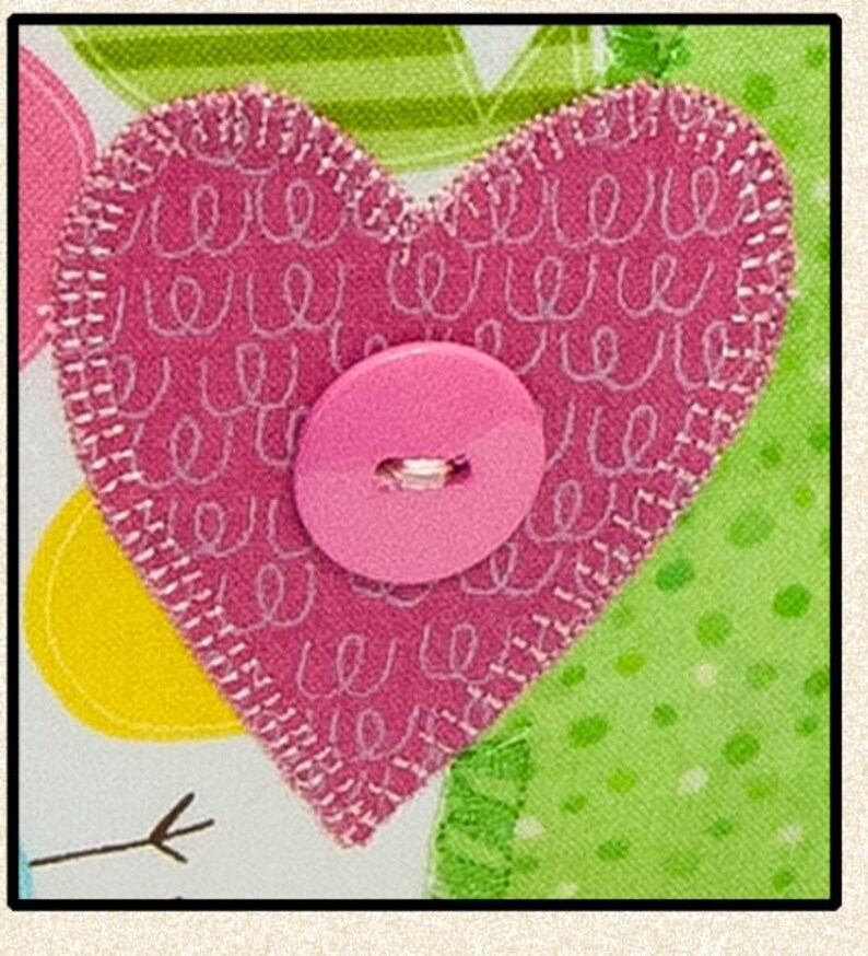 Pode incluir: Um cora&ccedil;&atilde;o de tecido rosa com bordas costuradas brancas e um bot&atilde;o rosa no centro. O cora&ccedil;&atilde;o &eacute; decorado com redemoinhos brancos e assenta sobre um fundo padronizado com detalhes verdes e amarelos. O design &eacute; alegre e artesanal.