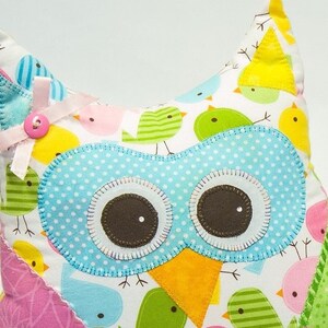 Pode incluir: Uma almofada colorida em forma de coruja com um design extravagante. A coruja apresenta um rosto azul claro com bolinhas, olhos castanhos grandes e um bico amarelo. A almofada &eacute; adornada com um la&ccedil;o e um bot&atilde;o rosa. O tecido de fundo &eacute; branco com v&aacute;rios p&aacute;ssaros coloridos.