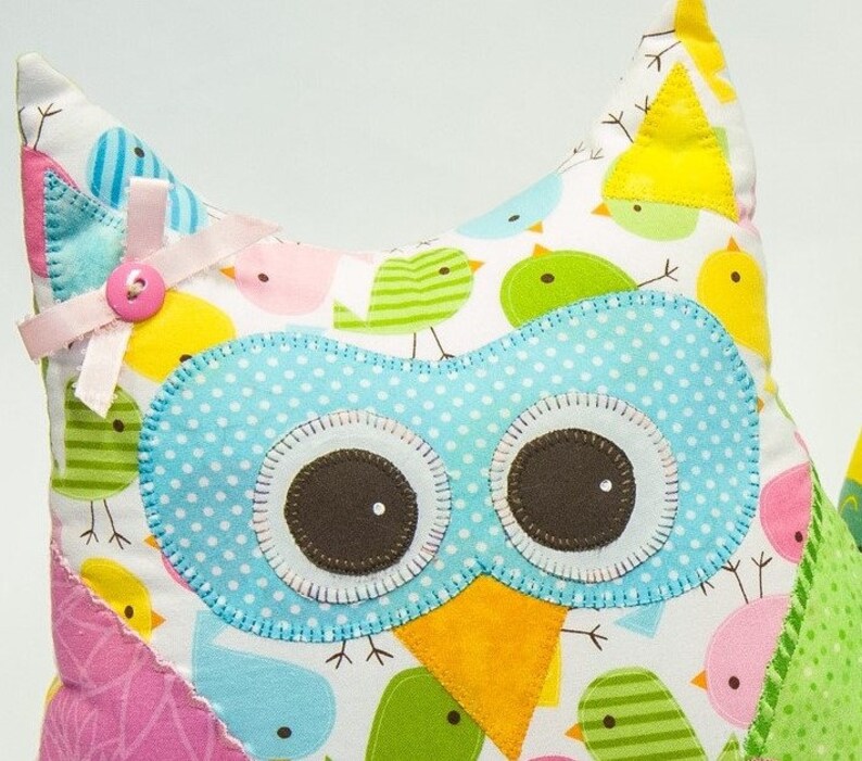 Pode incluir: Uma almofada colorida em forma de coruja com um design extravagante. A coruja apresenta um rosto azul claro com bolinhas, olhos castanhos grandes e um bico amarelo. A almofada &eacute; adornada com um la&ccedil;o e um bot&atilde;o rosa. O tecido de fundo &eacute; branco com v&aacute;rios p&aacute;ssaros coloridos.
