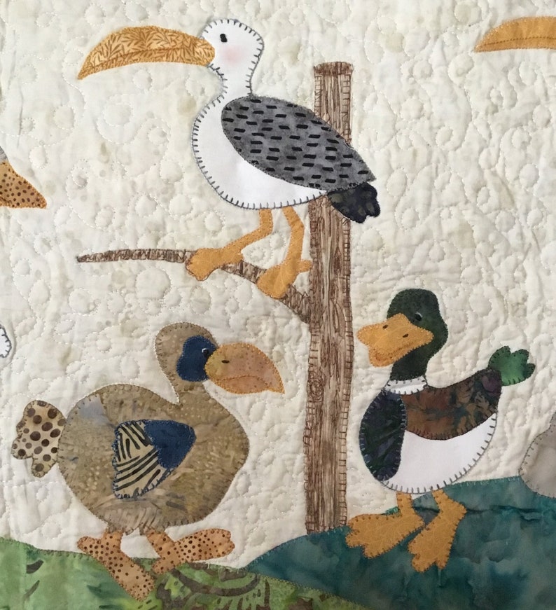 Puede incluir: Un tapiz de pared acolchado con p&aacute;jaros aplicados: una gaviota en una rama y dos patos. Las aves est&aacute;n hechas de telas estampadas en marr&oacute;n, azul, verde y blanco, sobre un fondo crema. El edred&oacute;n tiene un patr&oacute;n texturizado cosido.