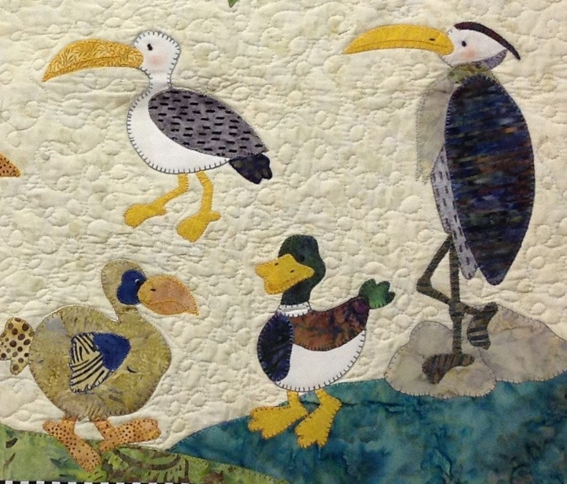 Puede incluir: Un textil acolchado con dise&ntilde;os caprichosos de aves. Las aves incluyen una gaviota, una garza y dos patos, cada uno con patrones de color y detalles &uacute;nicos. El fondo es de color beige claro con un patr&oacute;n acolchado, y las aves se colocan contra una escena de agua azul y verde.