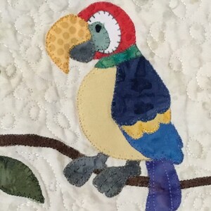 Puede incluir: Aplicaci&oacute;n de un loro colorido sobre un fondo acolchado. El loro presenta una cabeza roja, un pico amarillo y alas azules y amarillas. Est&aacute; posado en una rama marr&oacute;n con una hoja verde. El fondo es blanco con un patr&oacute;n acolchado.