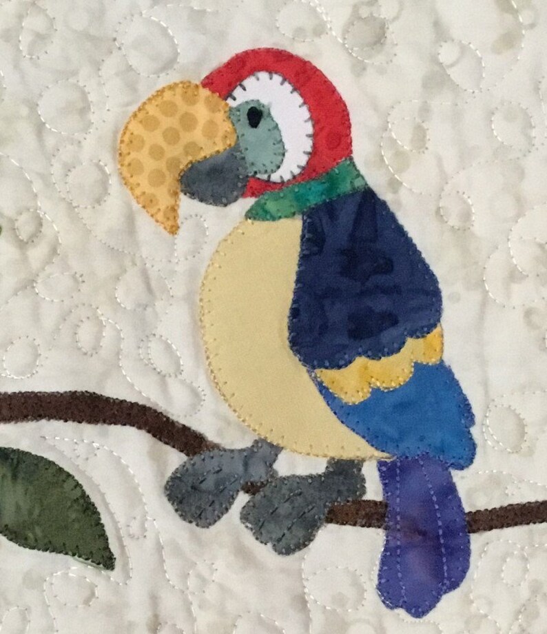 Puede incluir: Aplicaci&oacute;n de un loro colorido sobre un fondo acolchado. El loro presenta una cabeza roja, un pico amarillo y alas azules y amarillas. Est&aacute; posado en una rama marr&oacute;n con una hoja verde. El fondo es blanco con un patr&oacute;n acolchado.