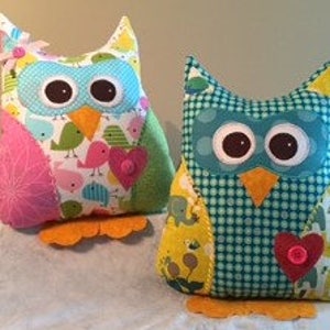 Pode incluir: Dois brinquedos de pel&uacute;cia em forma de coruja feitos &agrave; m&atilde;o. Uma coruja apresenta tecidos estampados rosa, azul e verde com enfeites de cora&ccedil;&atilde;o e flores. A outra coruja tem tecidos estampados azuis e amarelos com um cora&ccedil;&atilde;o e um bot&atilde;o. Ambas t&ecirc;m p&eacute;s laranja.