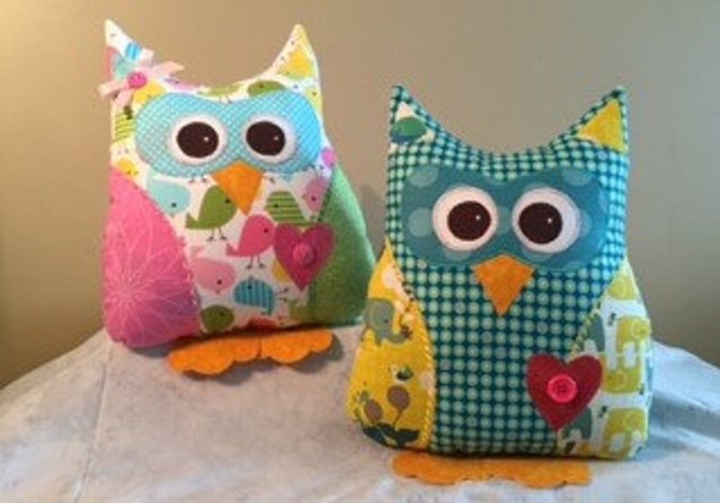 Pode incluir: Dois brinquedos de pel&uacute;cia em forma de coruja feitos &agrave; m&atilde;o. Uma coruja apresenta tecidos estampados rosa, azul e verde com enfeites de cora&ccedil;&atilde;o e flores. A outra coruja tem tecidos estampados azuis e amarelos com um cora&ccedil;&atilde;o e um bot&atilde;o. Ambas t&ecirc;m p&eacute;s laranja.