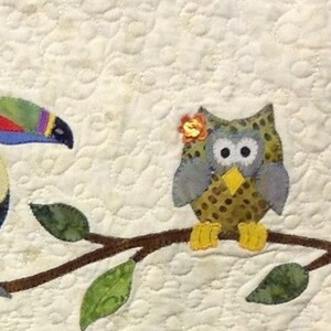 Puede incluir: Un tapiz de pared acolchado con un tuc&aacute;n y un b&uacute;ho posados en una rama con hojas verdes. El tuc&aacute;n tiene un pico colorido y el b&uacute;ho tiene una flor en la cabeza. El fondo es de color crema con un patr&oacute;n acolchado.