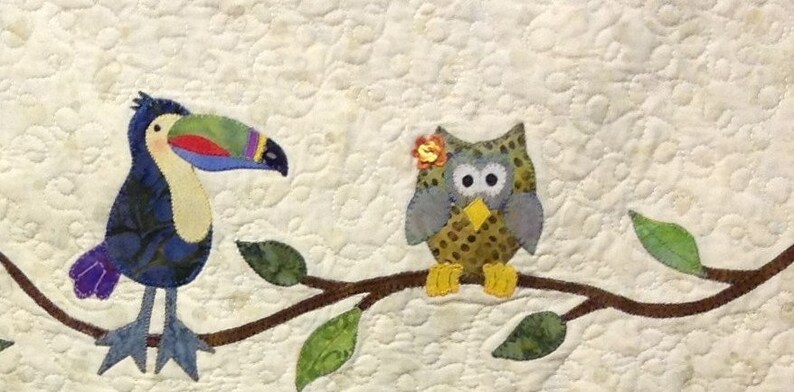 Puede incluir: Un tapiz de pared acolchado con un tuc&aacute;n y un b&uacute;ho posados en una rama con hojas verdes. El tuc&aacute;n tiene un pico colorido y el b&uacute;ho tiene una flor en la cabeza. El fondo es de color crema con un patr&oacute;n acolchado.