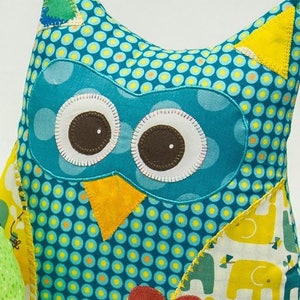 Pode incluir: Uma almofada artesanal em forma de coruja com um padr&atilde;o de bolinhas verde-azulado. A coruja tem olhos grandes e redondos com centros castanhos e contornos brancos. Apresenta orelhas triangulares amarelas, um bico amarelo e um cora&ccedil;&atilde;o vermelho com um bot&atilde;o rosa. As asas t&ecirc;m um padr&atilde;o de elefante amarelo.