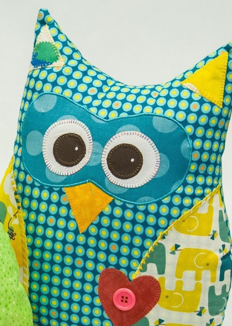 Pode incluir: Uma almofada artesanal em forma de coruja com um padr&atilde;o de bolinhas verde-azulado. A coruja tem olhos grandes e redondos com centros castanhos e contornos brancos. Apresenta orelhas triangulares amarelas, um bico amarelo e um cora&ccedil;&atilde;o vermelho com um bot&atilde;o rosa. As asas t&ecirc;m um padr&atilde;o de elefante amarelo.