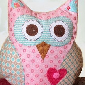 Pode incluir: Um brinquedo de pel&uacute;cia em forma de coruja rosa feito &agrave; m&atilde;o com olhos castanhos grandes e um patch em forma de cora&ccedil;&atilde;o. A coruja apresenta v&aacute;rios tecidos estampados em rosa, azul e castanho, com um detalhe de bot&atilde;o no cora&ccedil;&atilde;o. A coruja tem uma apar&ecirc;ncia artesanal e caprichosa.