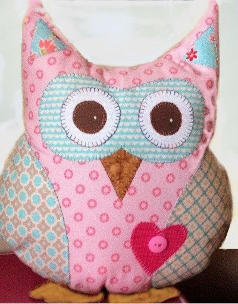 Pode incluir: Um brinquedo de pel&uacute;cia em forma de coruja rosa feito &agrave; m&atilde;o com olhos castanhos grandes e um patch em forma de cora&ccedil;&atilde;o. A coruja apresenta v&aacute;rios tecidos estampados em rosa, azul e castanho, com um detalhe de bot&atilde;o no cora&ccedil;&atilde;o. A coruja tem uma apar&ecirc;ncia artesanal e caprichosa.