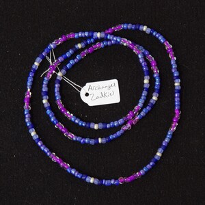 Puede incluir: Un collar de cuentas de m&uacute;ltiples hilos con cuentas de color morado, azul y blanco. El collar tiene una etiqueta que dice "Archangel Zadkiel".