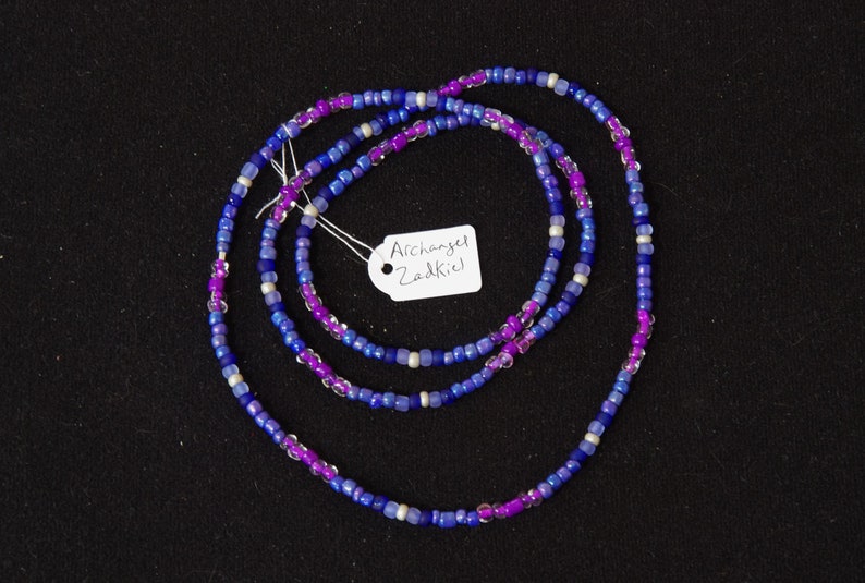 Puede incluir: Un collar de cuentas de m&uacute;ltiples hilos con cuentas de color morado, azul y blanco. El collar tiene una etiqueta que dice "Archangel Zadkiel".