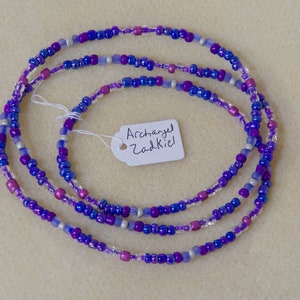 Puede incluir: Un collar de cuentas de m&uacute;ltiples hilos con cuentas de color morado, azul, plateado y transparente. Una etiqueta blanca con el texto "Archangel Zadkiel" est&aacute; unida al collar.