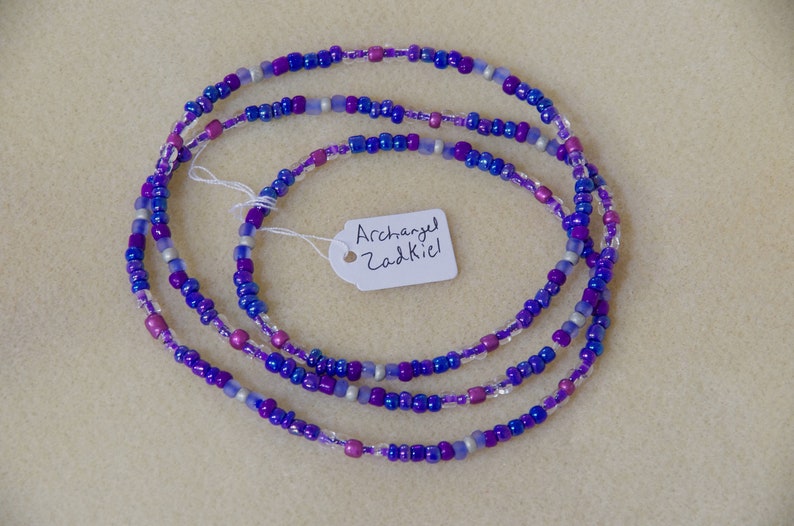 Puede incluir: Un collar de cuentas de m&uacute;ltiples hilos con cuentas de color morado, azul, plateado y transparente. Una etiqueta blanca con el texto "Archangel Zadkiel" est&aacute; unida al collar.