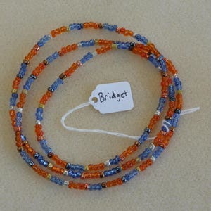 May include: A multi-strand beaded necklace with orange and blue beads. The necklace has a tag attached with the word "Bridget" written on it.