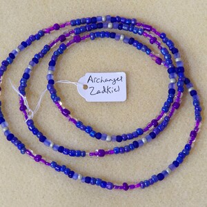 Puede incluir: Un collar de cuentas de m&uacute;ltiples hilos con cuentas de color morado, azul y plateado. El collar tiene una etiqueta adjunta que dice "Archangel Zadkiel".