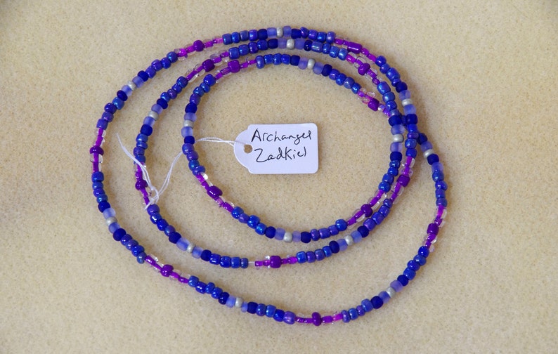 Puede incluir: Un collar de cuentas de m&uacute;ltiples hilos con cuentas de color morado, azul y plateado. El collar tiene una etiqueta adjunta que dice "Archangel Zadkiel".