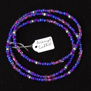 Puede incluir: Un brazalete de cuentas de tres hebras con cuentas moradas, azules y blancas. El brazalete tiene una etiqueta adjunta que dice "Arc&aacute;ngel Zadkiel".