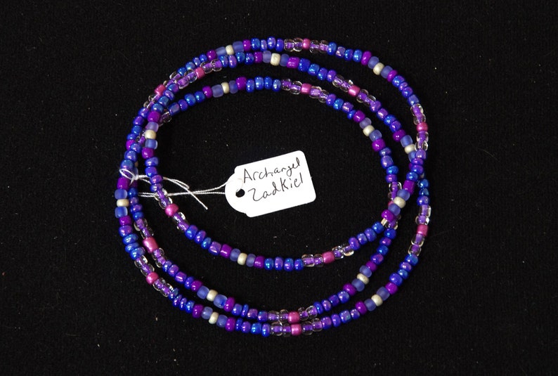 Puede incluir: Un brazalete de cuentas de tres hebras con cuentas moradas, azules y blancas. El brazalete tiene una etiqueta adjunta que dice "Arc&aacute;ngel Zadkiel".