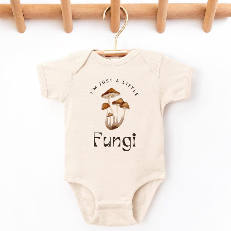 May include: A cream-colored baby onesie with short sleeves features a brown mushroom graphic and the text "I'm just a little Fungi".