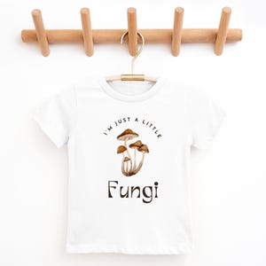 May include: White t-shirt with a brown mushroom graphic and the text "I'm just a little Fungi".