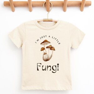 May include: A cream colored t-shirt with short sleeves features a graphic of mushrooms and the text "I'm just a little Fungi".