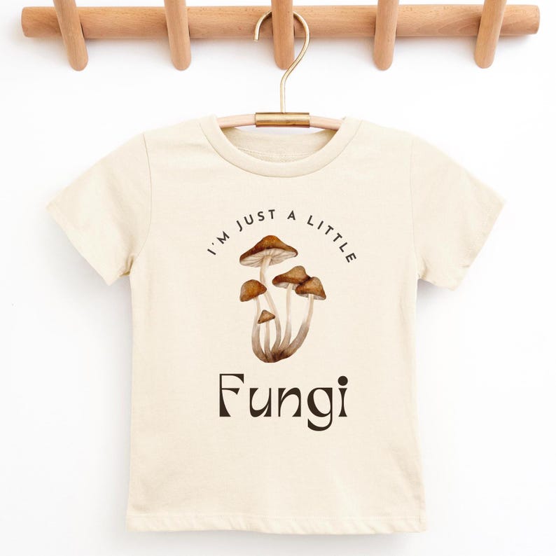 May include: A cream colored t-shirt with short sleeves features a graphic of mushrooms and the text "I'm just a little Fungi".