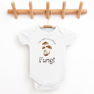 May include: White baby onesie with short sleeves featuring a brown mushroom illustration and the text "I'm just a little Fungi".