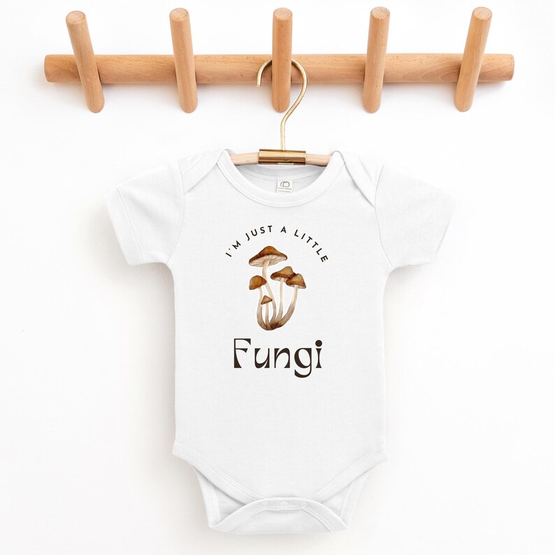 May include: White baby onesie with short sleeves featuring a brown mushroom illustration and the text "I'm just a little Fungi".