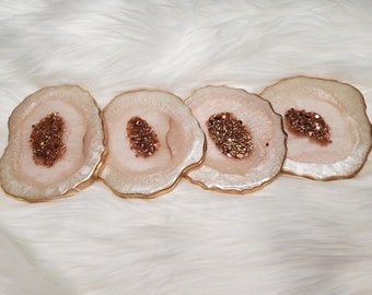 May include: Four resin coasters with a geode design. Each coaster has a light pink center with a sparkling, glittery brown center. The edges are outlined in gold, creating a luxurious look. The coasters are arranged on a white, fluffy surface.