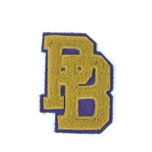 May include: A purple and gold felt letter B patch. The letter is outlined in purple and filled in with gold felt.