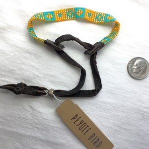 Pode incluir: Um bracelete de contas turquesa e amarelo com uma tira de couro marrom e uma conta de prata. O bracelete tem uma etiqueta que diz "Peyote Bird".